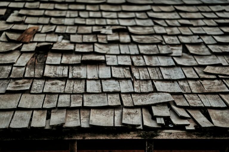 A roofing worker lifts shingles to inspect the wooden roof deck beneath an aging residential roof.